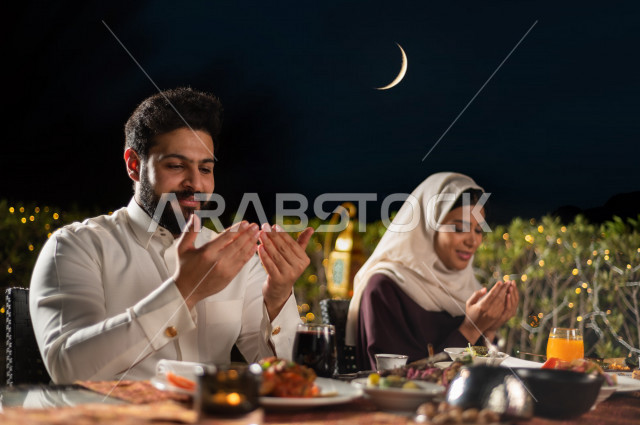 A Saudi Arabian couple at the breakfast table in the month of Ramadan, raising hands and praying to God with praise and thanksgiving, a trip full of varieties of delicious Saudi Arabian food, the month of Ramadan, Ramadan Kareem, Ramadan Lantern