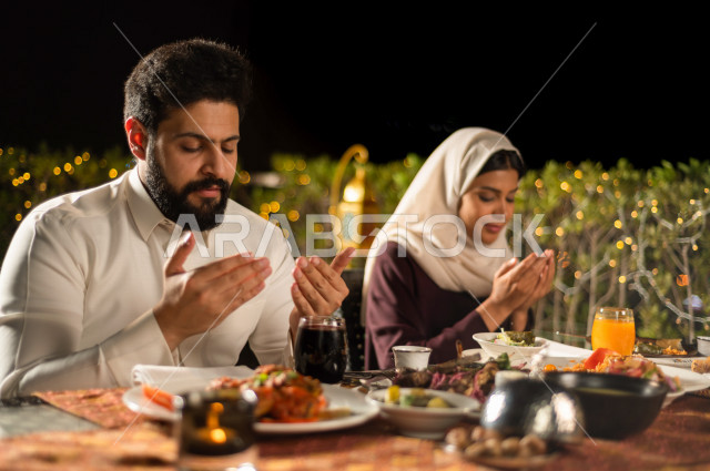 A Saudi Arabian couple at the breakfast table in the month of Ramadan, raising hands and praying to God with praise and thanksgiving, a trip full of varieties of delicious Saudi Arabian food, the month of Ramadan, Ramadan Kareem, Ramadan Lantern