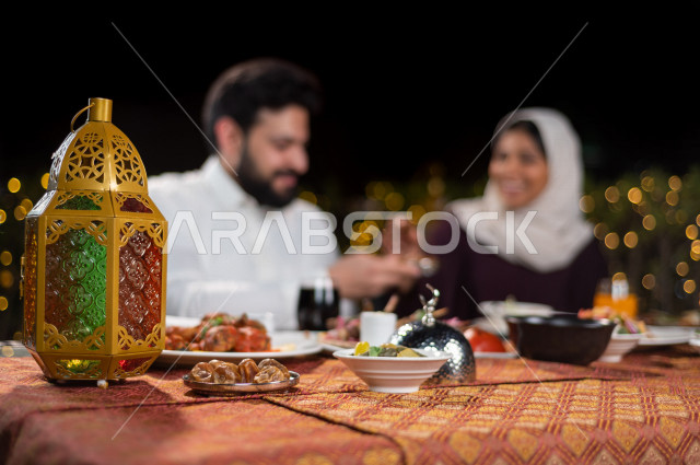زوجان سعوديان خليجيان على مائدة الافطار بشهررمضان ، مشاركة الاجواء الرمضانية الرائعة بالهواء الطلق ، سفرة مليئة بأصناف من المأكولات العربية السعودية اللذيذة ، شهر رمضان ، رمضان كريم ، فانوس رمضان