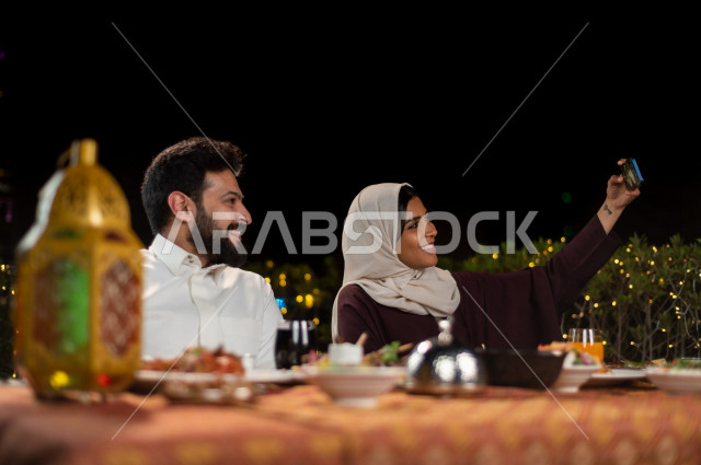 زوجان سعوديان خليجيان على مائدة الافطار بشهررمضان ، تصويرصورة سلفي من خلال الهاتف المحمول  ، مشاركة الاجواء الرمضانية ، سفرة مليئة بأصناف من المأكولات العربية السعودية اللذيذة ، شهر رمضان ، رمضان كريم ، فانوس رمضان