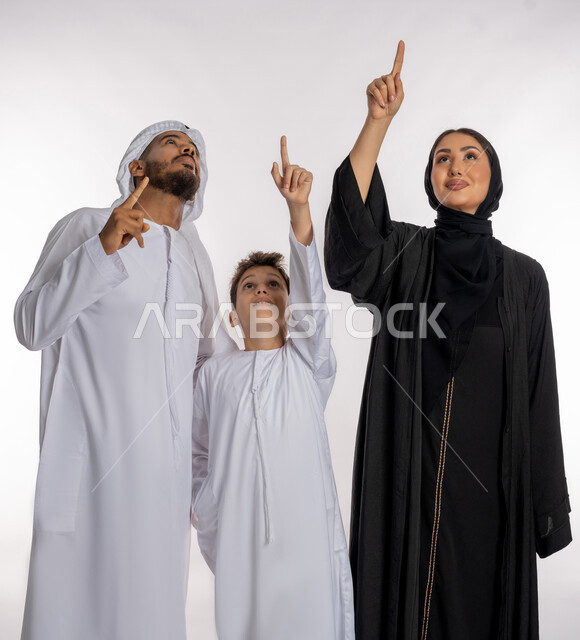 لمة العائلة الدافئة والترابط الأسري, قضاء الأوقات الممتعة برفقة الأسرة, بورتريه أب وأم عربيان خليجيان إماراتيان مع ولدهما الصغير, الاشارة إلى شيء ما بعيد والنظر إليه بإيماءات الاعجاب, مفهوم الأسرة الإماراتية السعيدة, خلفية بيضاء