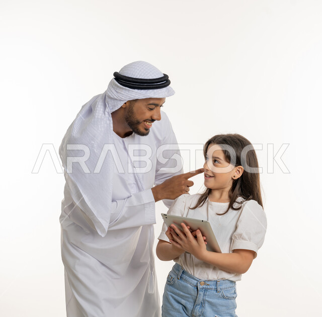 حب الأب لابنته الصغيرة, بورتريه لرجل عربي خليجي إماراتي يرتدي الكندورة والغترة ويقوم بمداعبة ابنته, استخدام التابلت في المكالمات والدردشات وتصفح الانترنت, الفتاة تنظر إلى والدها بإيماءات الحب والفرح, خلفية بيضاء