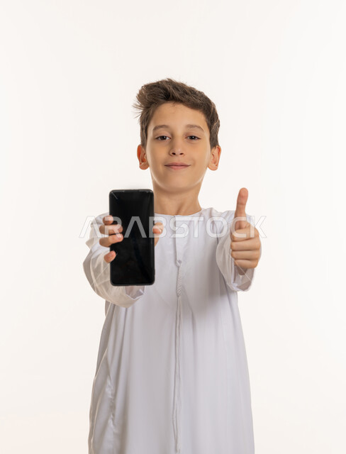 مواكبة التطور والتقدم في الامارات، عرض هاتف محمول بشاشة سوداء فارغة، دمج العلم مع التقنية والتكنولوجيا الحديثة، بورتريه مقرب لفتى عربي خليجي إماراتي يرتدي الكندورة الإماراتية ويرفع اصبعه بإيماءات الاعجاب, النظر إلى الكاميرا بسعادة، خلفية بيضاء