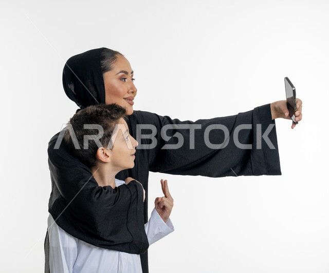 اهتمام الام بطفلها ورعايته, مفهوم الأمومة والحب والحنان, بورتريه لأم عربية خليجية إماراتية محجبة ترتدي العباءة تحضتن ابنها بين ذراعيها وتقوم باخذ صورة سيلفي لهما, التقاط الصور التذكارية الجميلة, خلفية بيضاء