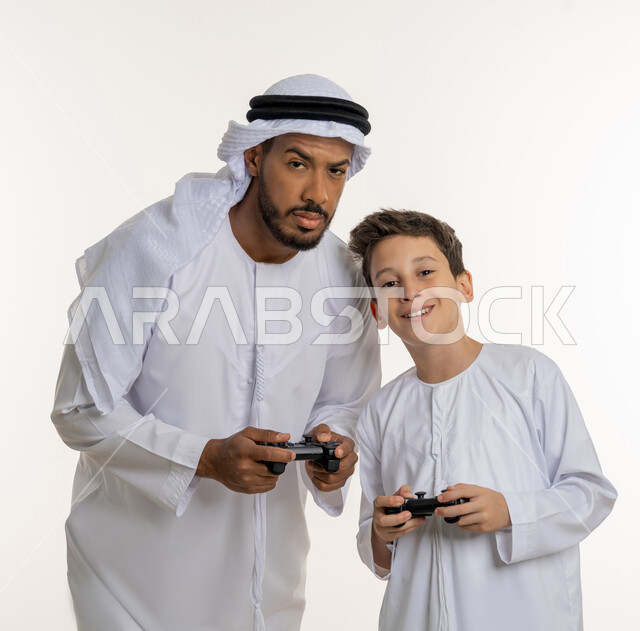 قضاء الأوقات العائلية الممتعة, بورتريه لأب عربي خليجي إماراتي برفقة ابنه الصغير يرتدون الكندورة الاماراتيه, النظر إلى الكاميرا بإيماءات التركيز والمتعة, والد يلعب مع ولده بالبلايستسشن، مفهوم الاسرة الإماراتية السعيدة، خلفية بيضاء