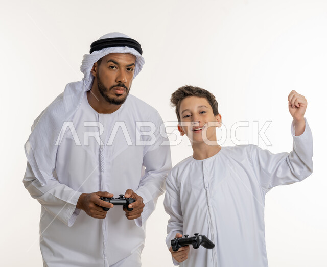 رفع اليد بتعبيرات النصر والفوز، قضاء الأوقات العائلية الممتعة, بورتريه لأب عربي خليجي إماراتي برفقة ابنه الصغير يرتدون الكندورة الاماراتيه, النظر إلى الكاميرا بإيماءات التركيز والمتعة, والد يلعب مع ولده بالبلايستسشن، مفهوم الاسرة الإماراتية السعيدة، خلفية بيضاء