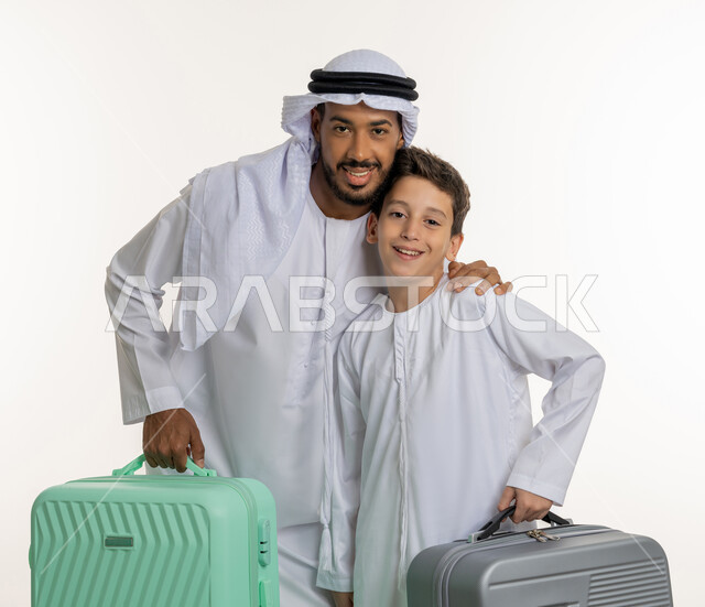 السفر وقضاء الإجازات برفقة الأبناء, بورتريه لعائلة عربية خليجية إماراتية تستعد للذهاب برحلة ممتعة, أب وابنه يمسكان بحقائب السفر وينظران إلى الكاميرا بإيماءات وجه تدل على السعادة، زي إماراتي تقليدي كندورة وغترة, سياحة و سفر في الامارات، خلفية بيضاء