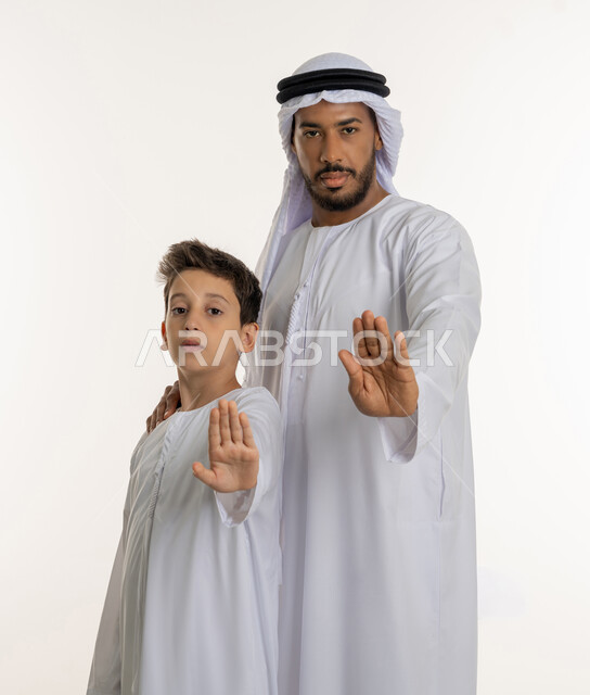 Hand gestures indicating the command to stop doing something and forbidding it, a child accompanied by his father, wearing the Emirati kandura and feeling annoyed, rejected and abstained, silent and stopping talking, a portrait of an Arab Emirati Gulf family raising their palms up, white background