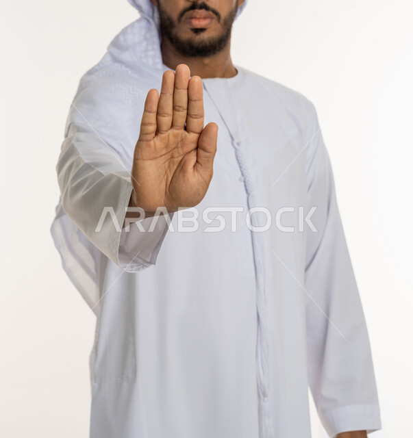 Raising the palm up with gestures indicating the command to stop doing something and forbidding it, a portrait of a Gulf Arab Kandora man feeling annoyed, rejected and abstained, silence, stopping talking, using sign language, white background.