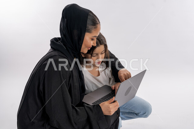 مساعدة الأبناء في حل الواجبات الدراسية، مفهوم التعلم عن بعد باستخدام الأجهزة التقنية الحديثة والتكنولوجيا، بورتريه أم عربية خليجية إماراتية محجبة تلبس العباءة وتجلس أمام الكومبيوتر المحمول مع ابنتها لمتابعة الدروس أونلاين، التعليم في الامارات، خلفية بيضاء