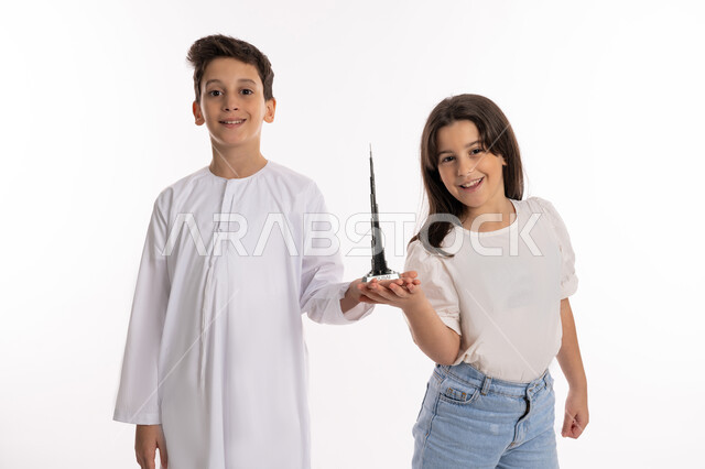 Family bonding and supporting sibling ties between children, a portrait of two Emirati Gulf Arab brothers holding a model of Burj Khalifa and looking at the camera with happy gestures, sharing recreational times between children, the concept of a happy Emirati family, white background
