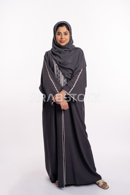 أزياء محجبات عصرية راقية، مفهوم الأناقة والاهتمام بالمظهر الخارجي، التقاط صورة شخصية للوثائق الرسمية، بورتريه لامرأة عربية خليجية إماراتية محجبة ترتدي العباءة الحديثة وتنظر الى الكاميرا بإيماءات السرور و الثقة بالنفس، صورة الجسم بالكامل، خلفية بيضاء