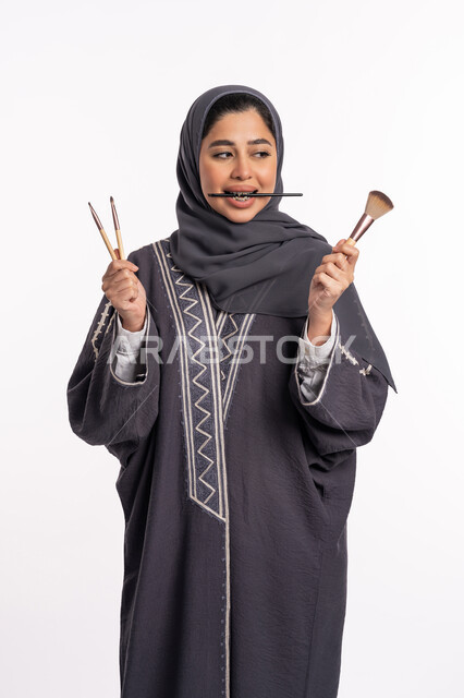 دورات تعليمية لمكياج تتوريال، مفهوم الإهتمام بالجمال والبشرة والمظهر الخارجي، خبيرة سعودية لمستحضرات التجميل النسائية، بورتريه لإمرأة عربية خليجية إماراتية ترتدي العباءة والحجاب تختار بين مجموعة من فرش الميكاب، خلفيه بيضاء