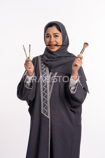 دورات تعليمية لمكياج تتوريال، مفهوم الإهتمام بالجمال والبشرة والمظهر الخارجي، خبيرة سعودية لمستحضرات التجميل النسائية، بورتريه لإمرأة عربية خليجية إماراتية ترتدي العباءة والحجاب تختار بين مجموعة من فرش الميكاب، خلفيه بيضاء