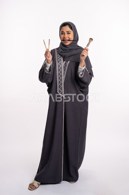 خبيرة سعودية لمستحضرات التجميل النسائية، دورات تعليمية لمكياج تتوريال، مفهوم الإهتمام بالجمال والبشرة والمظهر الخارجي، بورتريه لإمرأة عربية خليجية إماراتية ترتدي العباءة والحجاب تختار بين مجموعة من فرش الميكاب، صورة الجسم بالكامل، خلفيه بيضاء