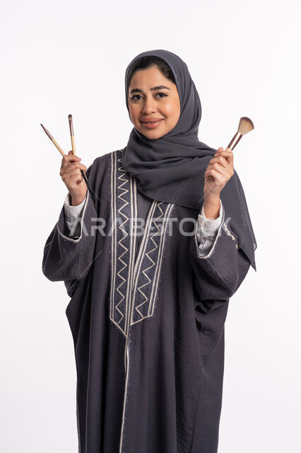 دورات تعليمية لمكياج تتوريال، مفهوم الإهتمام بالجمال والبشرة والمظهر الخارجي، خبيرة سعودية لمستحضرات التجميل النسائية، بورتريه لإمرأة عربية خليجية إماراتية ترتدي العباءة والحجاب تختار بين مجموعة من فرش الميكاب، خلفيه بيضاء