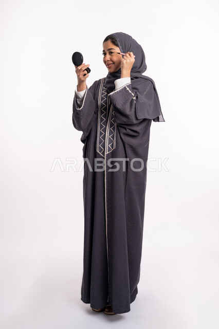 Using eyeliner to highlight the beauty of the eyes, paying attention to beauty and external appearance, educational courses for editorial makeup, a Saudi expert in women’s cosmetics, a close-up portrait of an Arab Gulf-Emirati woman wearing the abaya and hijab, holding a black eyeliner pencil, full body portrait, white background
