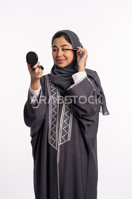 استخدام الماسكرا لإبراز جمال العينين، الإهتمام بالجمال والمظهر الخارجي، دورات تعليمية لمكياج تتوريال, خبيرة سعودية لمستحضرات التجميل النسائية، بورتريه مقرب لإمرأة عربية خليجية إماراتية ترتدي العباءة والحجاب تقوم بوضع الميكاب، خلفيه بيضاء