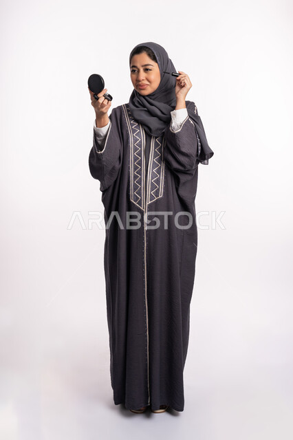 Using mascara to highlight the beauty of the eyes, paying attention to beauty and external appearance, educational courses for editorial makeup, a Saudi expert in women’s cosmetics, a close-up portrait of an Arab Gulf-Emirati woman wearing the abaya and hijab, applying makeup, full body portrait, white background