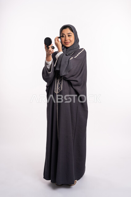 استخدام الماسكرا لإبراز جمال العينين، الإهتمام بالجمال والمظهر الخارجي، دورات تعليمية لمكياج تتوريال, خبيرة سعودية لمستحضرات التجميل النسائية، بورتريه لإمرأة عربية خليجية إماراتية ترتدي العباءة والحجاب تقوم بوضع الميكاب، صورة الجسم بالكامل، خلفيه بيضاء