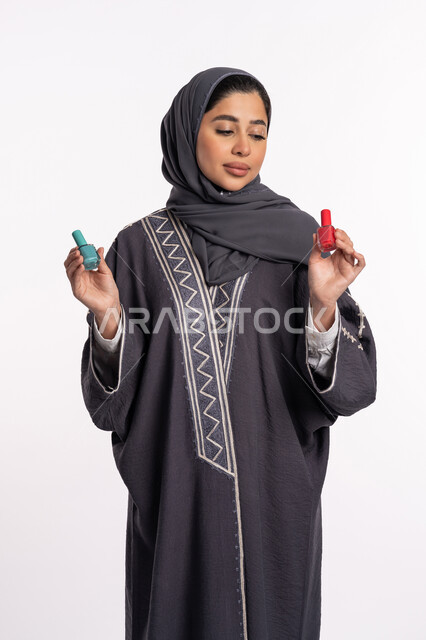 الحيرة في اختيار لون طلاء الأظافر، خبيرة سعودية لمستحضرات التجميل النسائية، صالون تجميلي نسائي، بورتريه لإمرأة عربية خليجية إماراتية ترتدي العباءة والحجاب تحمل بيدها المانيكير، مفهوم الإهتمام بالجمال والمظهر الخارجي، خلفية بيضاء
