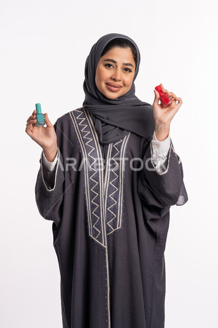 مفهوم الإهتمام بالجمال والمظهر الخارجي، صالون تجميلي نسائي، الحيرة في اختيار لون طلاء الأظافر، خبيرة سعودية لمستحضرات التجميل النسائية، بورتريه لإمرأة عربية خليجية إماراتية ترتدي العباءة والحجاب تحمل بيدها المانيكير، خلفية بيضاء