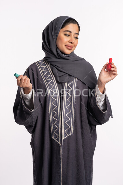 الحيرة في اختيار لون طلاء الأظافر، خبيرة سعودية لمستحضرات التجميل النسائية، صالون تجميلي نسائي، بورتريه لإمرأة عربية خليجية إماراتية ترتدي العباءة والحجاب تحمل بيدها المانيكير، مفهوم الإهتمام بالجمال والمظهر الخارجي، خلفية بيضاء