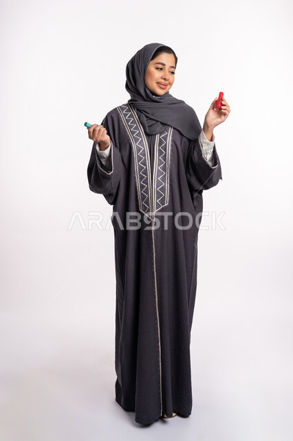 صالون تجميلي نسائي، الحيرة في اختيار لون طلاء الأظافر، خبيرة سعودية لمستحضرات التجميل النسائية، بورتريه لإمرأة عربية خليجية إماراتية ترتدي العباءة والحجاب تحمل بيدها المانيكير، مفهوم الإهتمام بالجمال والمظهر الخارجي، صورة الجسم بالكامل، خلفية بيضاء