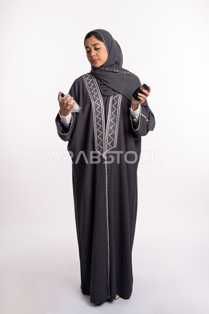 الحيرة في اختيار الرائحة، منتجات العطور النسائيه، بورتريه لإمرأة عربية خليجية إماراتية ترتدي العباءة والحجاب تقوم برش العطر، الأناقة والإهتمام بالمظهر الخارجي، استخدام الزيوت العطرية الطبيعية، التطيب والتعطر بالعود العطري، صورة الجسم بالكامل، خلفية بيضاء