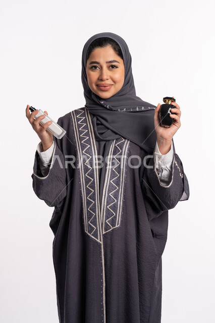 The use of natural essential oils, perfuming and perfuming with aromatic oud, confusion in choosing a scent, women’s perfume products, a portrait of an Arab Gulf Emirati woman wearing an abaya and hijab, spraying perfume, elegance and concern for external appearance, a white background.