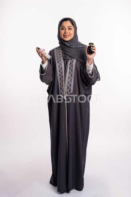 Elegance and attention to external appearance, perfuming and perfuming with aromatic oud, using natural essential oils, confusion in choosing a scent, women’s perfume products, a portrait of an Arab Gulf Emirati woman wearing an abaya and hijab, spraying perfume, full body portrait, white background