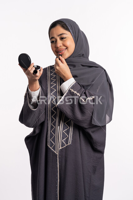 دورات تعليمية لمكياج تتوريال، مفهوم الإهتمام بالجمال والبشرة والمظهر الخارجي, خبيرة سعودية لمستحضرات التجميل النسائية، بورتريه لإمرأة عربية خليجية إماراتية ترتدي العباءة والحجاب تحمل مرآة تقوم بوضع احمر الشفاه، خلفيه بيضاء