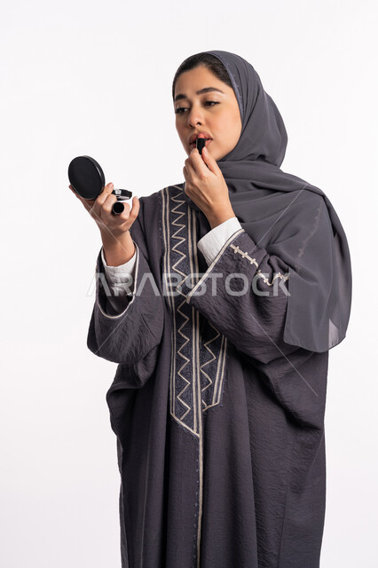 دورات تعليمية لمكياج تتوريال، مفهوم الإهتمام بالجمال والبشرة والمظهر الخارجي, خبيرة سعودية لمستحضرات التجميل النسائية، بورتريه لإمرأة عربية خليجية إماراتية ترتدي العباءة والحجاب تحمل مرآة تقوم بوضع احمر الشفاه، خلفيه بيضاء