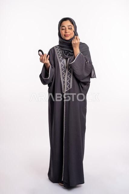Applying shadows to highlight the beauty of the eyes, a Saudi expert in women’s cosmetics, a portrait of an Arab Gulf Emirati woman wearing the abaya and hijab, applying eyeshadow, educational courses for portrait makeup, the concept of caring for beauty, skin, and external appearance, full body image, white background
