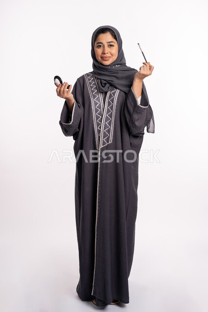 وضع الظلال لإبراز جمال العيون، خبيرة سعودية لمستحضرات التجميل النسائية، بورتريه لإمرأة عربية خليجية إماراتية ترتدي العباءة والحجاب تقوم بوضع الايشادو، دورات تعليمية لمكياج تتوريال، مفهوم الإهتمام بالجمال والبشرة والمظهر الخارجي, صورة الجسم بالكامل، خلفيه بيضاء