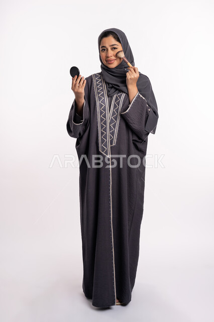 استخدام احمر الخدود لتوريد الوجه، دورات تعليمية لمكياج تتوريال، خبيرة سعودية لمستحضرات التجميل، بورتريه لإمرأة عربية خليجية إماراتية ترتدي العباءة والحجاب تقوم بوضع البلاشر، الإهتمام بالجمال والمظهر الخارجي, صورة الجسم بالكامل، خلفيه بيضاء