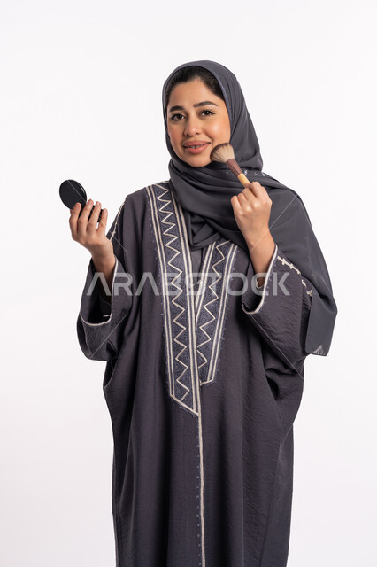 استخدام احمر الخدود لتوريد الوجه، دورات تعليمية لمكياج تتوريال، خبيرة سعودية لمستحضرات التجميل، بورتريه لإمرأة عربية خليجية إماراتية ترتدي العباءة والحجاب تقوم بوضع البلاشر، الإهتمام بالجمال والمظهر الخارجي, خلفيه بيضاء