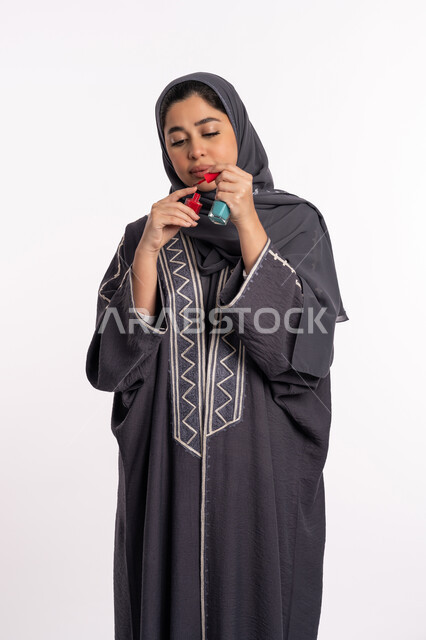 استخدام لون لطلاء الأظافر، خبيرة سعودية لمستحضرات التجميل النسائية، بورتريه لإمرأة عربية خليجية إماراتية ترتدي العباءة والحجاب تحمل بيدها المانيكير، مفهوم الإهتمام بالجمال والمظهر الخارجي، صالون تجميلي نسائي، خلفية بيضاء