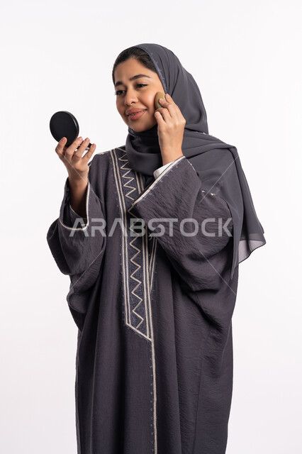 Paying attention to beauty and external appearance, applying foundation to hide skin blemishes, educational courses for cosmetic make-up, a Saudi cosmetics expert, a portrait of an Arab Gulf-Emirati woman wearing an abaya and hijab, holding a beauty blender and a mirror in her hand, a white background.
