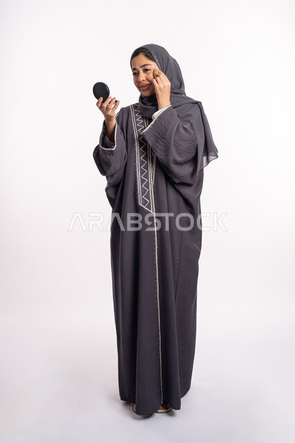 وضع كريم الأساس لإخفاء عيوب البشرة، الإهتمام بالجمال والمظهر الخارجي, دورات تعليمية لمكياج تتوريال، خبيرة سعودية لمستحضرات التجميل، بورتريه لإمرأة عربية خليجية إماراتية ترتدي العباءة والحجاب تحمل بيدها البيوتي بلندر والمرآة، صورة الجسم بالكامل، خلفيه بيضاء