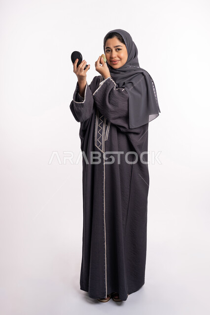 وضع كريم الأساس لإخفاء عيوب البشرة، الإهتمام بالجمال والمظهر الخارجي, دورات تعليمية لمكياج تتوريال، خبيرة سعودية لمستحضرات التجميل، بورتريه لإمرأة عربية خليجية إماراتية ترتدي العباءة والحجاب تحمل بيدها البيوتي بلندر والمرآة، صورة الجسم بالكامل، خلفيه بيضاء