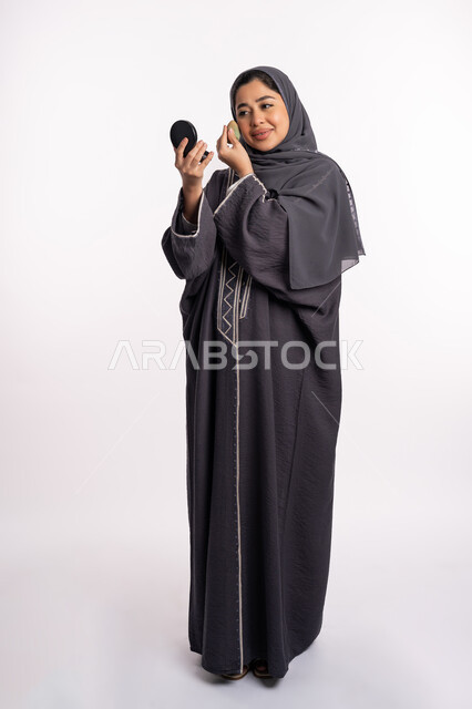 وضع كريم الأساس لإخفاء عيوب البشرة، الإهتمام بالجمال والمظهر الخارجي, دورات تعليمية لمكياج تتوريال، خبيرة سعودية لمستحضرات التجميل، بورتريه لإمرأة عربية خليجية إماراتية ترتدي العباءة والحجاب تحمل بيدها البيوتي بلندر والمرآة، صورة الجسم بالكامل، خلفيه بيضاء