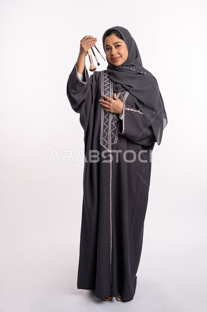 مفهوم الإهتمام بالجمال والبشرة والمظهر الخارجي, دورات تعليمية لمكياج تتوريال، خبيرة سعودية لمستحضرات التجميل النسائية، بورتريه لإمرأة عربية خليجية إماراتية ترتدي العباءة والحجاب تختار بين مجموعة من فرش الميكاب، صورة الجسم بالكامل، خلفيه بيضاء