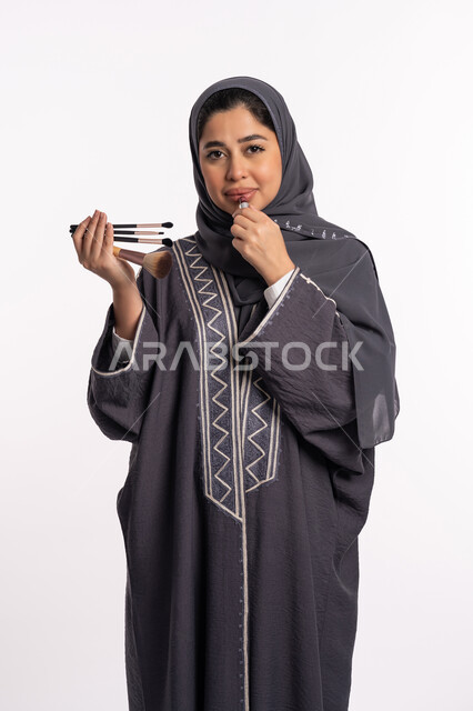 وضع احمر الشفاه، دورات تعليمية لمكياج تتوريال، خبيرة سعودية لمستحضرات التجميل النسائية، مفهوم الإهتمام بالجمال والبشرة والمظهر الخارجي, بورتريه لإمرأة عربية خليجية إماراتية ترتدي العباءة والحجاب تختار بين مجموعة من فرش الميكاب، خلفيه بيضاء