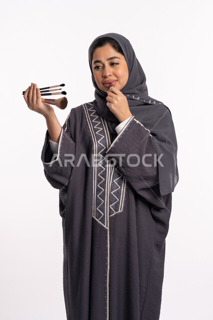 Applying lipstick, educational courses for tutorial makeup, a Saudi expert in women’s cosmetics, the concept of caring for beauty, skin, and external appearance, a portrait of an Arab Gulf Emirati woman wearing an abaya and hijab, choosing between a group of makeup brushes, a white background.