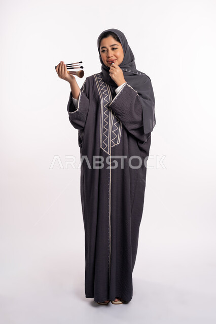 دورات تعليمية لمكياج تتوريال، خبيرة سعودية لمستحضرات التجميل النسائية، وضع احمر الشفاه، مفهوم الإهتمام بالجمال والبشرة والمظهر الخارجي, بورتريه لإمرأة عربية خليجية إماراتية ترتدي العباءة والحجاب تختار بين مجموعة من فرش الميكاب، صورة الجسم بالكامل، خلفيه بيضاء