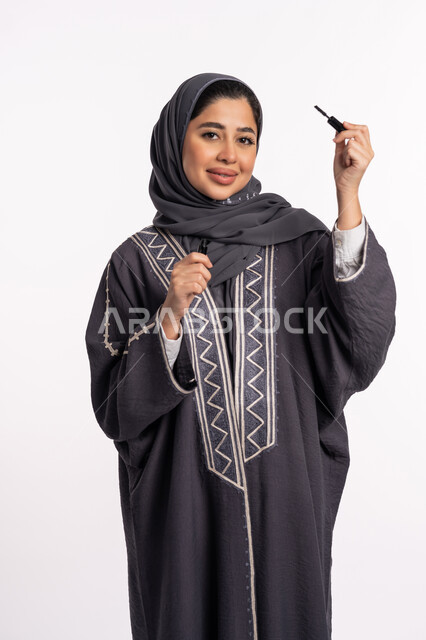 Highlighting the beauty of the eyes, the concept of caring for beauty, skin, and external appearance, educational courses for makeup, a Saudi expert in women’s cosmetics, a close-up portrait of an Arab Gulf Emirati woman wearing an abaya and hijab, holding a black kohl pencil, a white background.