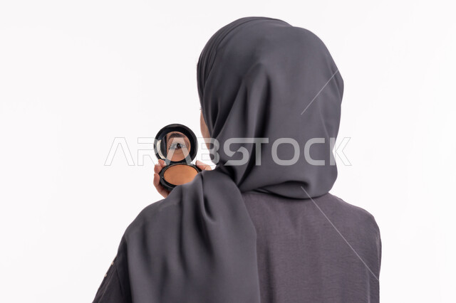 مفهوم الإهتمام بالجمال والبشرة والمظهر الخارجي، دورات تعليمية لمكياج تتوريال, خبيرة سعودية لمستحضرات التجميل النسائية، بورتريه مقرب من الخلف لإمرأة عربية خليجية إماراتية ترتدي العباءة والحجاب تحمل مرآة تقوم بوضع مورد الخدود، خلفيه بيضاء