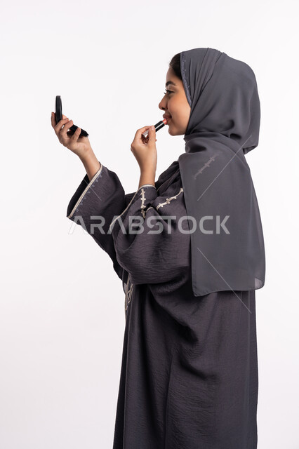 Tutorial makeup courses, the concept of caring for beauty, skin, and external appearance, a Saudi expert in women’s cosmetics, a portrait of an Arab Gulf-Emirati woman wearing an abaya and hijab, holding a mirror applying lipstick, white background.