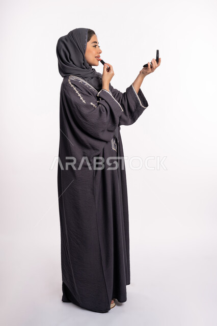 مفهوم الإهتمام بالجمال والبشرة والمظهر الخارجي، دورات تعليمية لمكياج تتوريال, خبيرة سعودية لمستحضرات التجميل النسائية، بورتريه لإمرأة عربية خليجية إماراتية ترتدي العباءة والحجاب تحمل مرآة تقوم بوضع احمر الشفاه، صورة الجسم بالطول الكامل، خلفيه بيضاء